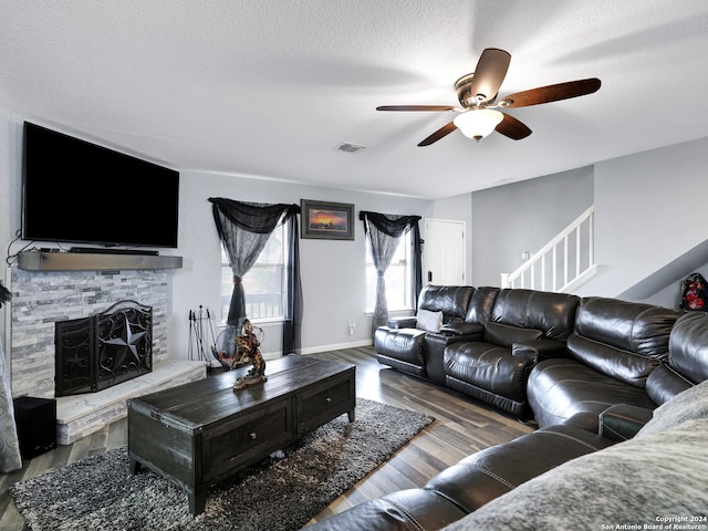 living room with ceiling fan, a textured ceiling, wood-type flooring, and a stone fireplace