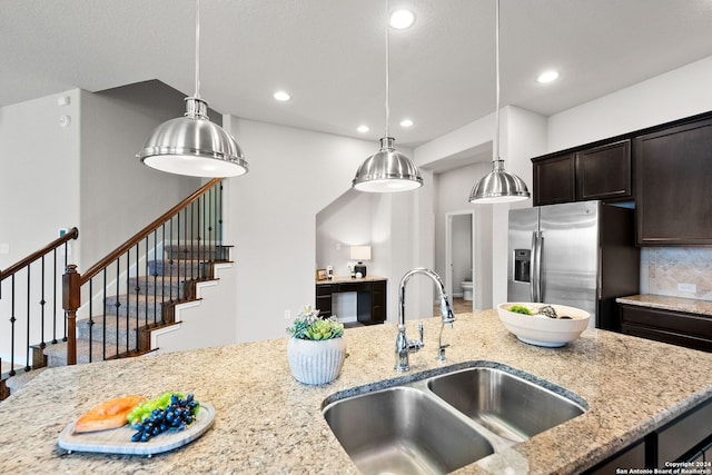 kitchen with dark brown cabinetry, light stone counters, a sink, stainless steel fridge, and decorative light fixtures
