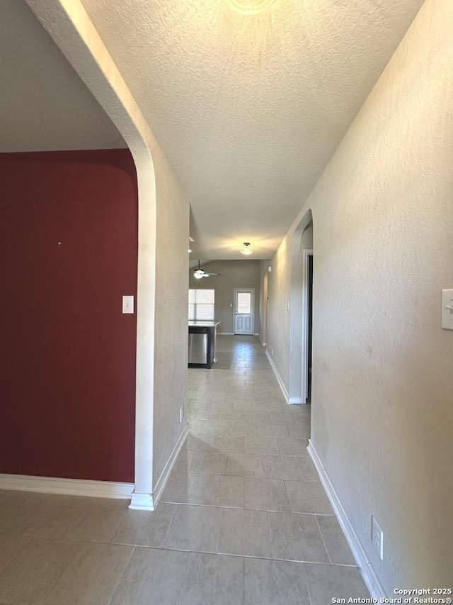 corridor featuring arched walkways, a textured wall, a textured ceiling, baseboards, and tile patterned floors