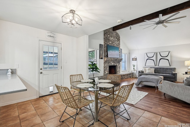 dining room with light wood-type flooring, ceiling fan with notable chandelier, vaulted ceiling with beams, and a brick fireplace