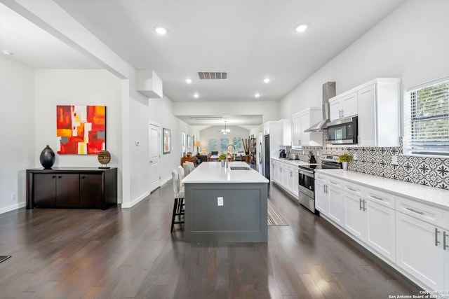 kitchen featuring wall chimney range hood, a breakfast bar, white cabinetry, stainless steel appliances, and an island with sink