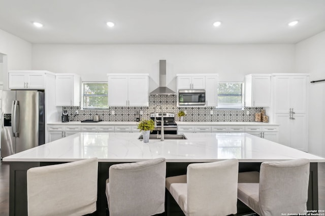 kitchen with white cabinetry, a large island with sink, light stone counters, stainless steel appliances, and wall chimney range hood