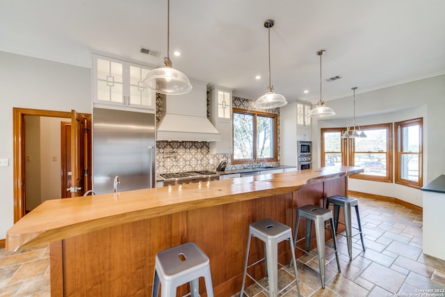 kitchen featuring built in appliances, custom range hood, white cabinetry, and a breakfast bar