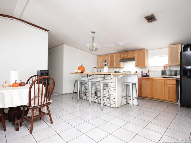 kitchen with a notable chandelier, under cabinet range hood, black refrigerator with ice dispenser, stainless steel microwave, and a kitchen bar