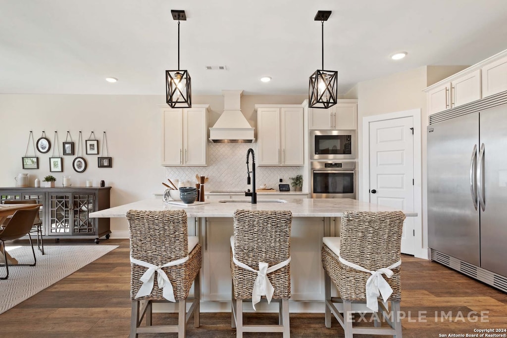 kitchen featuring a kitchen island with sink, built in appliances, light stone counters, dark wood-type flooring, and custom range hood