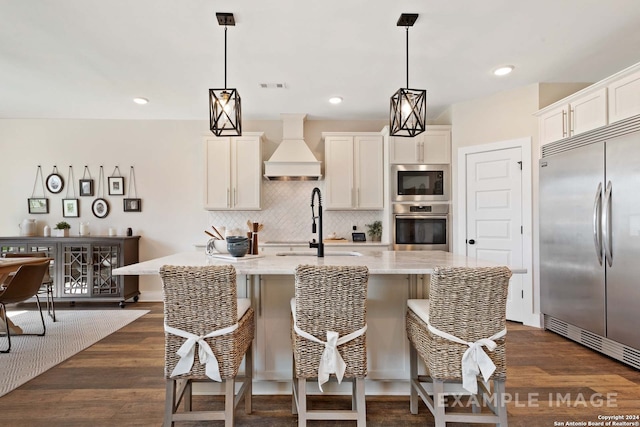 kitchen featuring a kitchen island with sink, built in appliances, light stone counters, dark wood-type flooring, and custom range hood