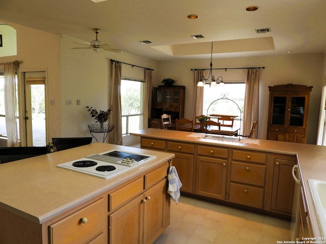 kitchen featuring a raised ceiling, white electric stovetop, a center island with sink, pendant lighting, and ceiling fan