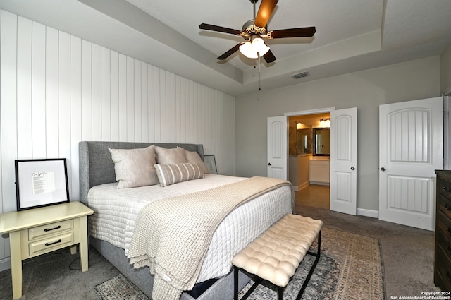 bedroom featuring a tray ceiling, dark carpet, ensuite bathroom, and ceiling fan