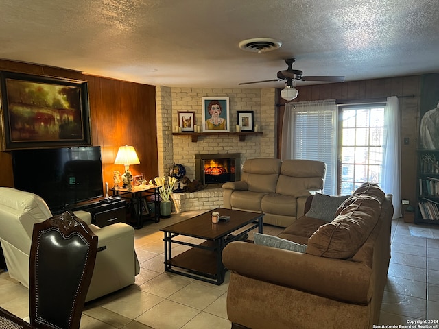 tiled living room with a textured ceiling, ceiling fan, wood walls, and a brick fireplace