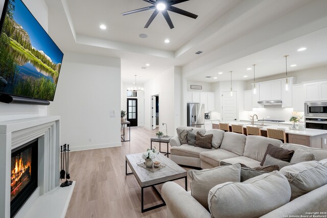 living room with sink, ceiling fan with notable chandelier, light wood-type flooring, and a tray ceiling