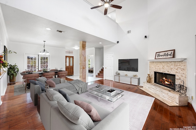 living room featuring dark wood-type flooring, a high ceiling, ceiling fan with notable chandelier, a stone fireplace, and ornamental molding