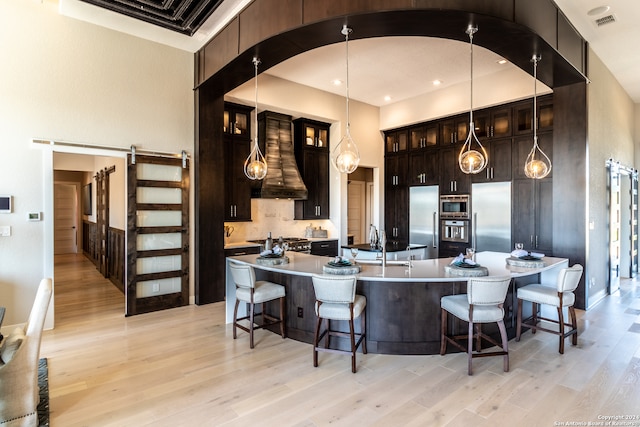 kitchen featuring built in appliances, a barn door, light wood-type flooring, decorative light fixtures, and custom range hood