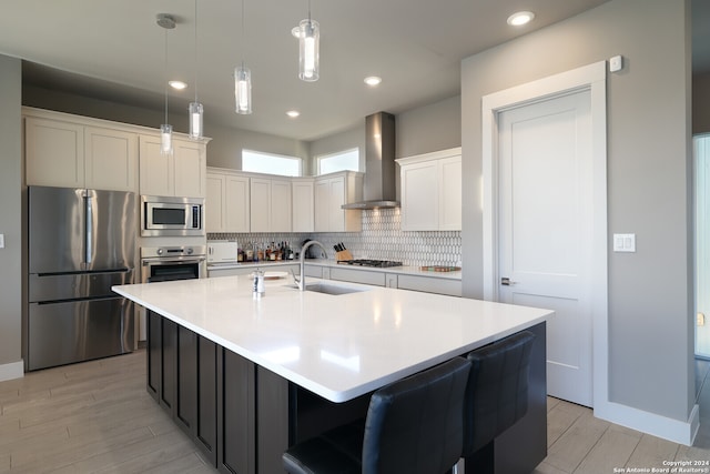 kitchen featuring stainless steel appliances, light hardwood / wood-style floors, sink, wall chimney range hood, and an island with sink