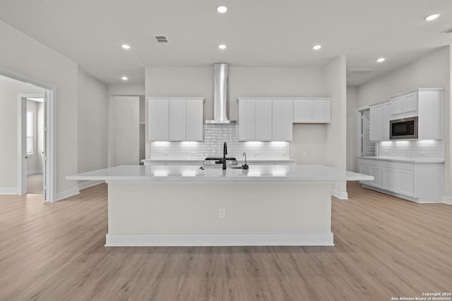 kitchen with light hardwood / wood-style flooring, a kitchen island with sink, stainless steel microwave, white cabinets, and wall chimney exhaust hood