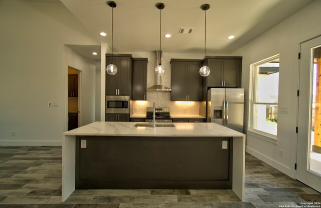 kitchen featuring an island with sink, backsplash, wall chimney range hood, stainless steel appliances, and light stone countertops