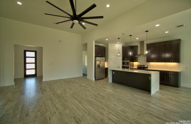 kitchen featuring light hardwood / wood-style floors, a kitchen island with sink, wall chimney exhaust hood, stainless steel appliances, and decorative light fixtures