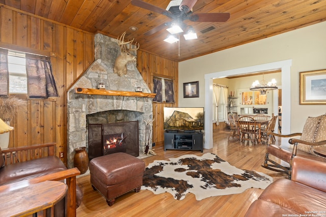 living room with ceiling fan with notable chandelier, wood-type flooring, wooden walls, and a stone fireplace