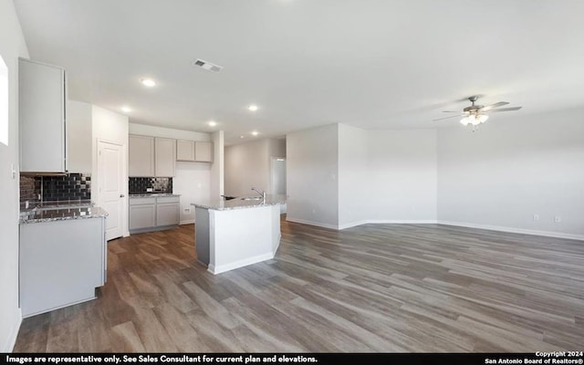 kitchen featuring light stone counters, an island with sink, dark wood-type flooring, ceiling fan, and decorative backsplash