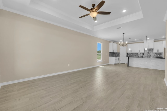 unfurnished living room with light wood-type flooring, ceiling fan with notable chandelier, crown molding, sink, and a tray ceiling