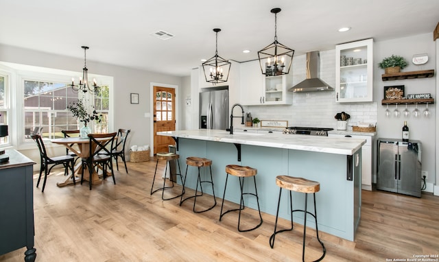 kitchen with an inviting chandelier, stainless steel appliances, white cabinetry, wall chimney exhaust hood, and a kitchen island with sink