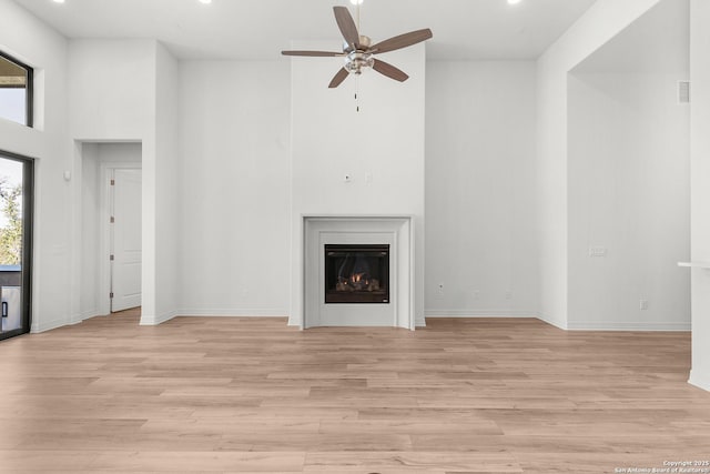 unfurnished living room with baseboards, ceiling fan, light wood-style flooring, a warm lit fireplace, and a high ceiling