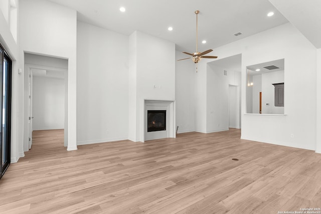 unfurnished living room with recessed lighting, light wood-style floors, a towering ceiling, a glass covered fireplace, and a ceiling fan