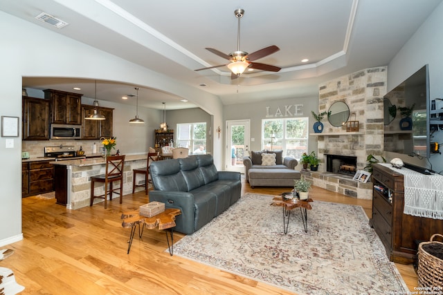 living room featuring a raised ceiling, a stone fireplace, ceiling fan, and light hardwood / wood-style floors