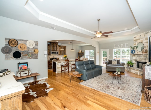 living room with a stone fireplace, ceiling fan, a raised ceiling, and light hardwood / wood-style flooring