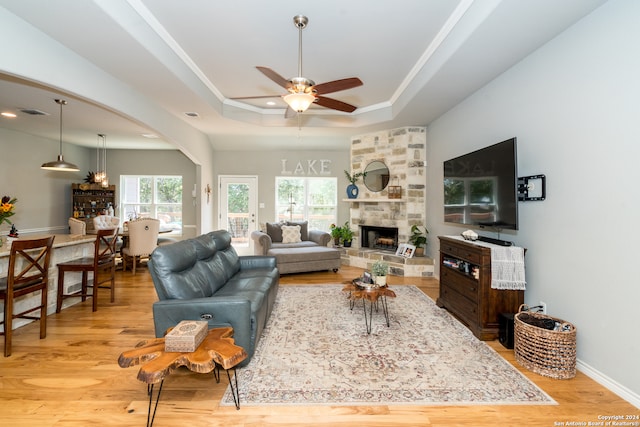 living room with light hardwood / wood-style flooring, ceiling fan, a raised ceiling, and a stone fireplace