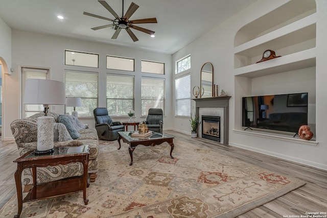 living room featuring baseboards, a tiled fireplace, a ceiling fan, built in features, and wood finished floors