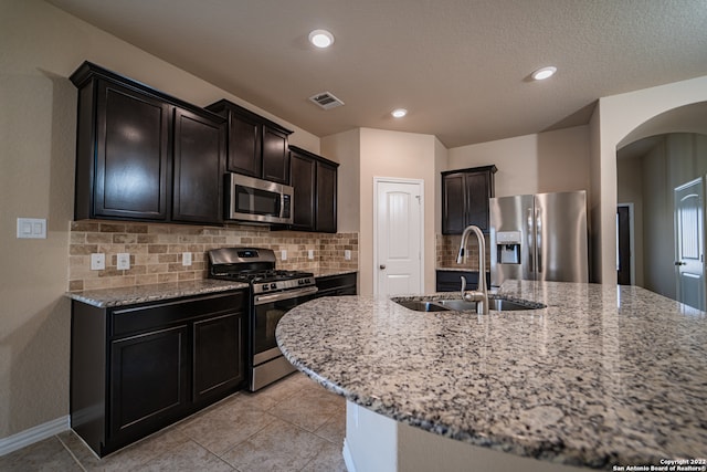 kitchen featuring backsplash, light stone counters, an island with sink, sink, and appliances with stainless steel finishes