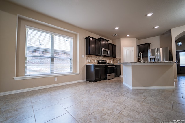 kitchen with a kitchen island with sink, backsplash, light tile patterned floors, stainless steel appliances, and light stone counters