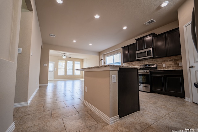 kitchen featuring stainless steel appliances, ceiling fan, decorative backsplash, a kitchen island with sink, and a textured ceiling