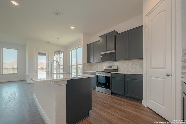 kitchen featuring dark wood-type flooring, stainless steel range oven, an island with sink, decorative light fixtures, and decorative backsplash