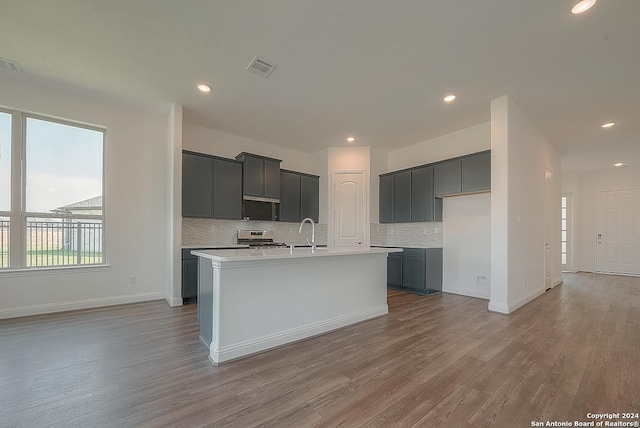 kitchen with sink, backsplash, a center island with sink, stainless steel stove, and light wood-type flooring