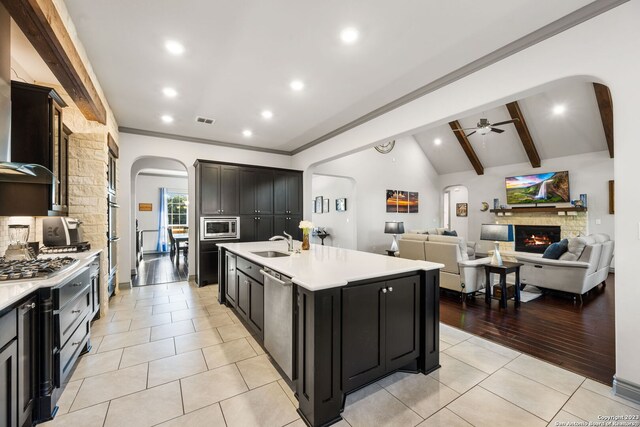 kitchen with light hardwood / wood-style floors, a stone fireplace, ceiling fan, a kitchen island with sink, and vaulted ceiling with beams