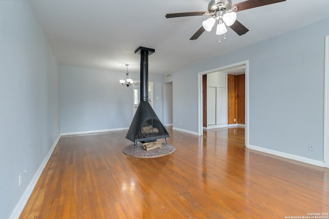 empty room with ceiling fan with notable chandelier, wood-type flooring, and a wood stove