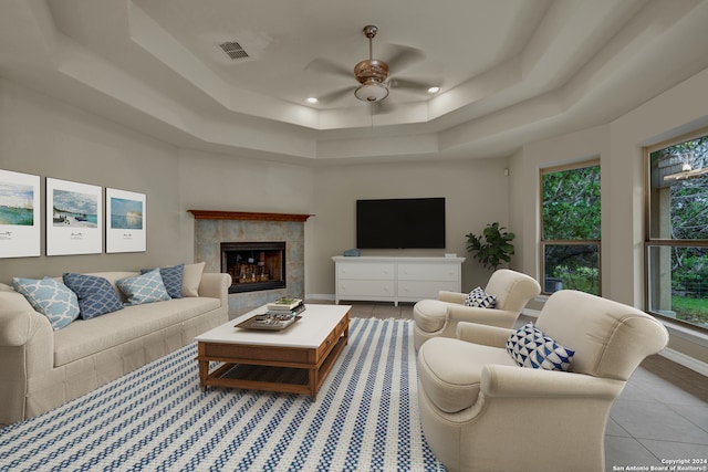 living room featuring a tiled fireplace, light tile patterned flooring, a tray ceiling, and ceiling fan