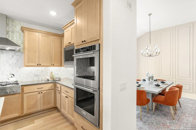 kitchen with wall chimney range hood, an inviting chandelier, hanging light fixtures, appliances with stainless steel finishes, and light hardwood / wood-style floors