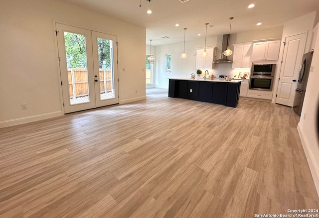 kitchen with wall chimney exhaust hood, pendant lighting, a kitchen island with sink, light wood-type flooring, and white cabinets