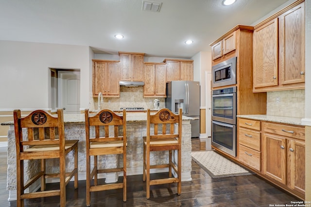 kitchen with appliances with stainless steel finishes, light stone counters, dark hardwood / wood-style flooring, a kitchen bar, and decorative backsplash