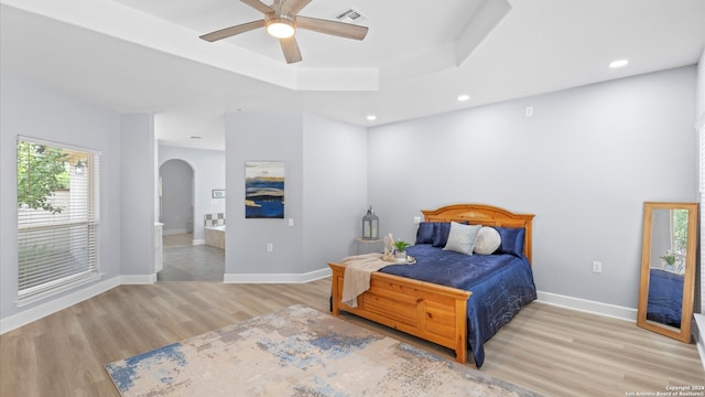 bedroom featuring light wood-type flooring, a tray ceiling, ceiling fan, and ensuite bathroom