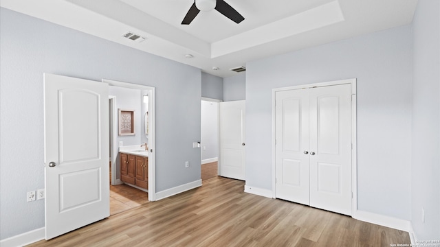 unfurnished bedroom with ceiling fan, a closet, a tray ceiling, light wood-type flooring, and ensuite bath