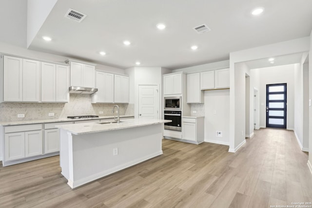 kitchen with sink, stainless steel appliances, white cabinetry, light stone counters, and an island with sink