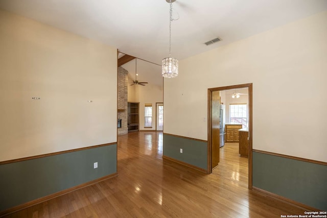 empty room with high vaulted ceiling, ceiling fan with notable chandelier, a fireplace, and light hardwood / wood-style flooring