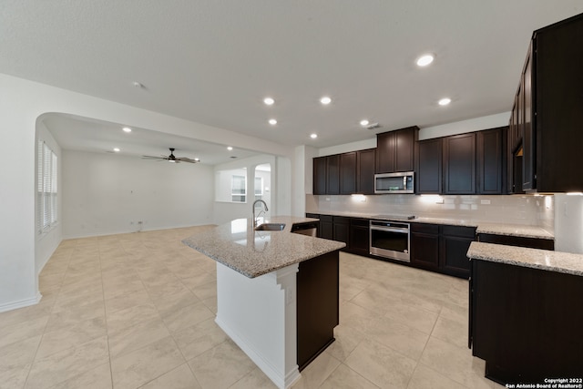 kitchen with appliances with stainless steel finishes, light stone counters, sink, ceiling fan, and a center island with sink