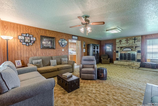 carpeted living room featuring a stone fireplace, wooden walls, ceiling fan, and a textured ceiling