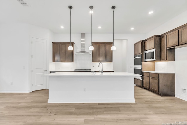 kitchen with stainless steel microwave, a kitchen island with sink, wall chimney range hood, sink, and hanging light fixtures