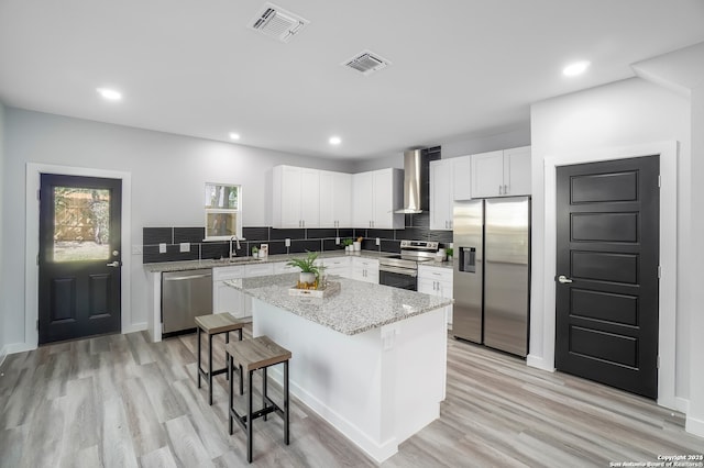 kitchen featuring white cabinets, wall chimney exhaust hood, stainless steel appliances, a center island, and light hardwood / wood-style floors