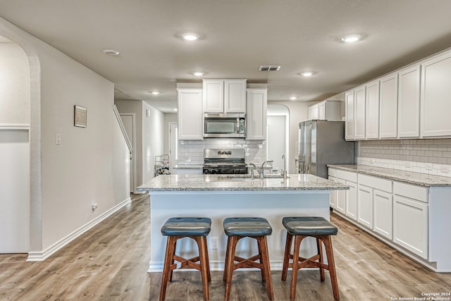 kitchen featuring light stone counters, light hardwood / wood-style floors, white cabinets, a center island with sink, and appliances with stainless steel finishes